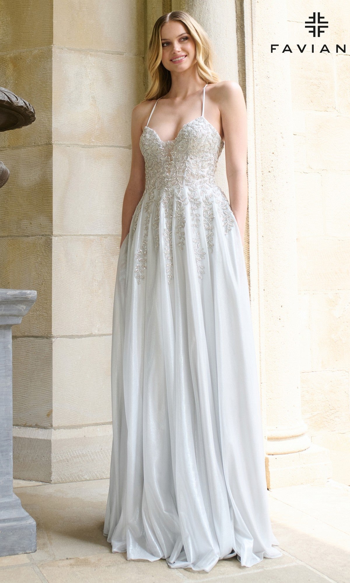 A woman wearing the Faviana 11449 long prom dress, a silver embellished A-line gown with thin straps and a fitted beaded bodice, stands before a stone wall.